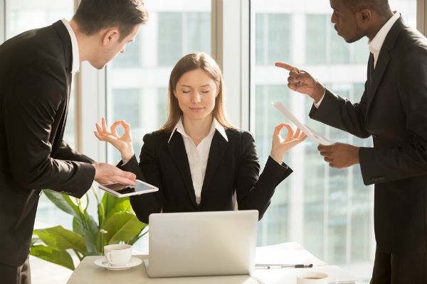 woman meditating in office while men yell at her | 26 Ways to Make Practicing Mindfulness Part of Your Day
