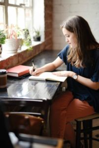 woman writing in notebook at coffee shop | Can Staying Positive Increase Your Productivity? https://positiveroutines.com/staying-positive-increase-productivity/