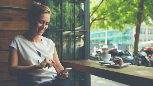 woman in cafe smiling at smartphone | 75 Great Apps That Will Change Your Life https://positiveroutines.com/great-apps-for-change/ 
