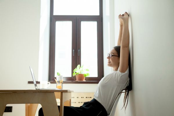 young woman stretching at desk near window | 59 Work Tips to Be Better At Your Job  https://positiveroutines.com/best-work-tips/