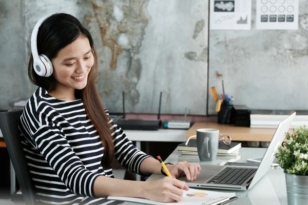 asian woman listening to music smiling at work | Can You Use Music For Productivity Gains? https://positiveroutines.com/music-for-productivity/