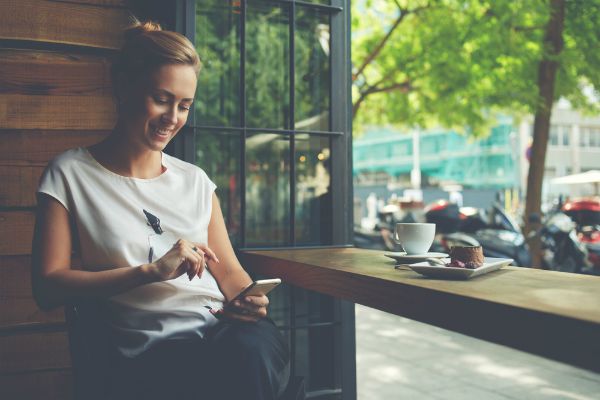 woman sitting in coffee shop smiling at phone | 83 Secrets To Make You Happier At Work https://positiveroutines.com/happier-at-work/