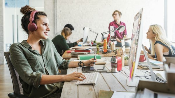 woman with headphones on smiling while working at open office | 83 Secrets To Make You Happier At Work https://positiveroutines.com/happier-at-work/