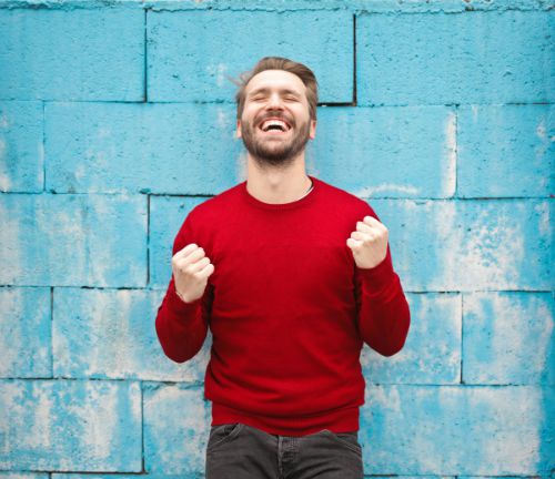 man in red sweater smiling with eyes closed against blue brick wall | 7 Surprising Ways to Be Successful at Work https://positiveroutines.com/successful-at-work/