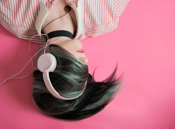 woman listening to white headphones with hair over her face against pink background | What Are Binaural Beats + Can They Boost Productivity? https://positiveroutines.com/what-are-binaural-beats/ ‎