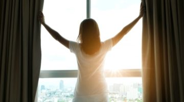 woman opening curtains to sunrise | A Healthy Morning Routine For The Best 2019 https://positiveroutines.com/healthy-morning-routine/