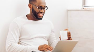 male working on laptop with mug in hand smiling | The Best Tips for Working Remotely When You're Outgoing, Anxious, and More https://positiveroutines.com/tips-for-working-remotely/