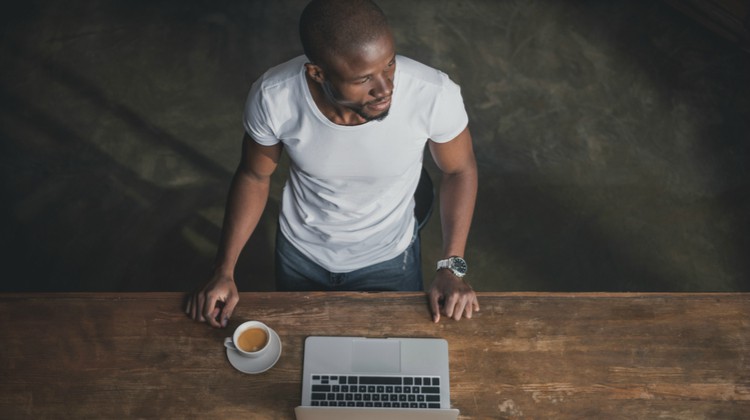 man standing at wooden table in front of laptop and coffee looking toward sunlight | Make These 5 Tiny Tweaks to Have A Great Morning https://positiveroutines.com/great-morning-tips/