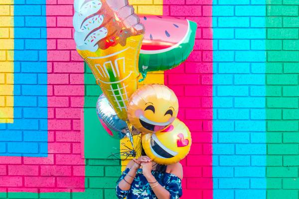 woman holding bouquet of happy emoji ballons standing in front of colorful brick wall | 7 Easy Ways to Build a More Positive Mindset https://positiveroutines.com/positive-mindset/