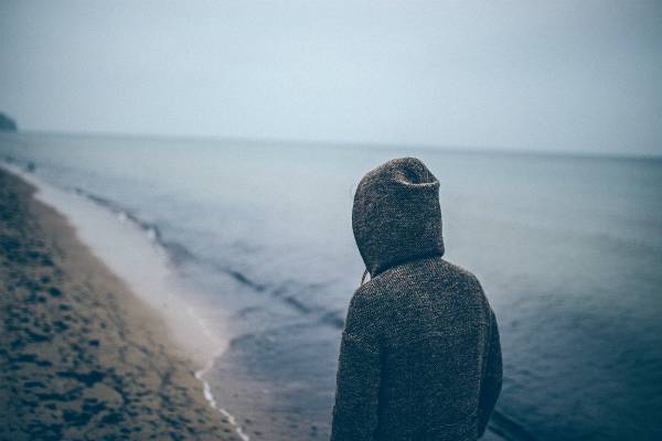 lonely person in hooded sweatshirt walking along beach | The Happiest Country in the World This Year is... https://positiveroutines.com/happiest-country-in-the-world/