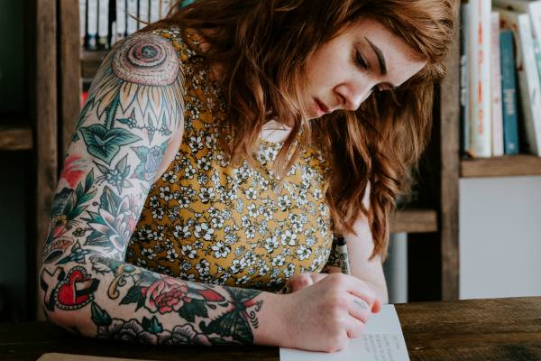 tattooed woman writing in card | 13 Gratitude Journal Prompts to Try This International Day of Happiness https://positiveroutines.com/gratitude-journal-prompts/