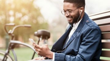 happy-male-on-park-bench-wearing-earbuds | 13 Ways to be Happier This Summer https://positiveroutines.com/be-happier-this-summer/