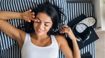 happy woman laying in bed beside headphones and sleep mask | 7 Sleep Podcasts to Help You Have the Best Rest Ever https://positiveroutines.com/sleep-podcasts/
